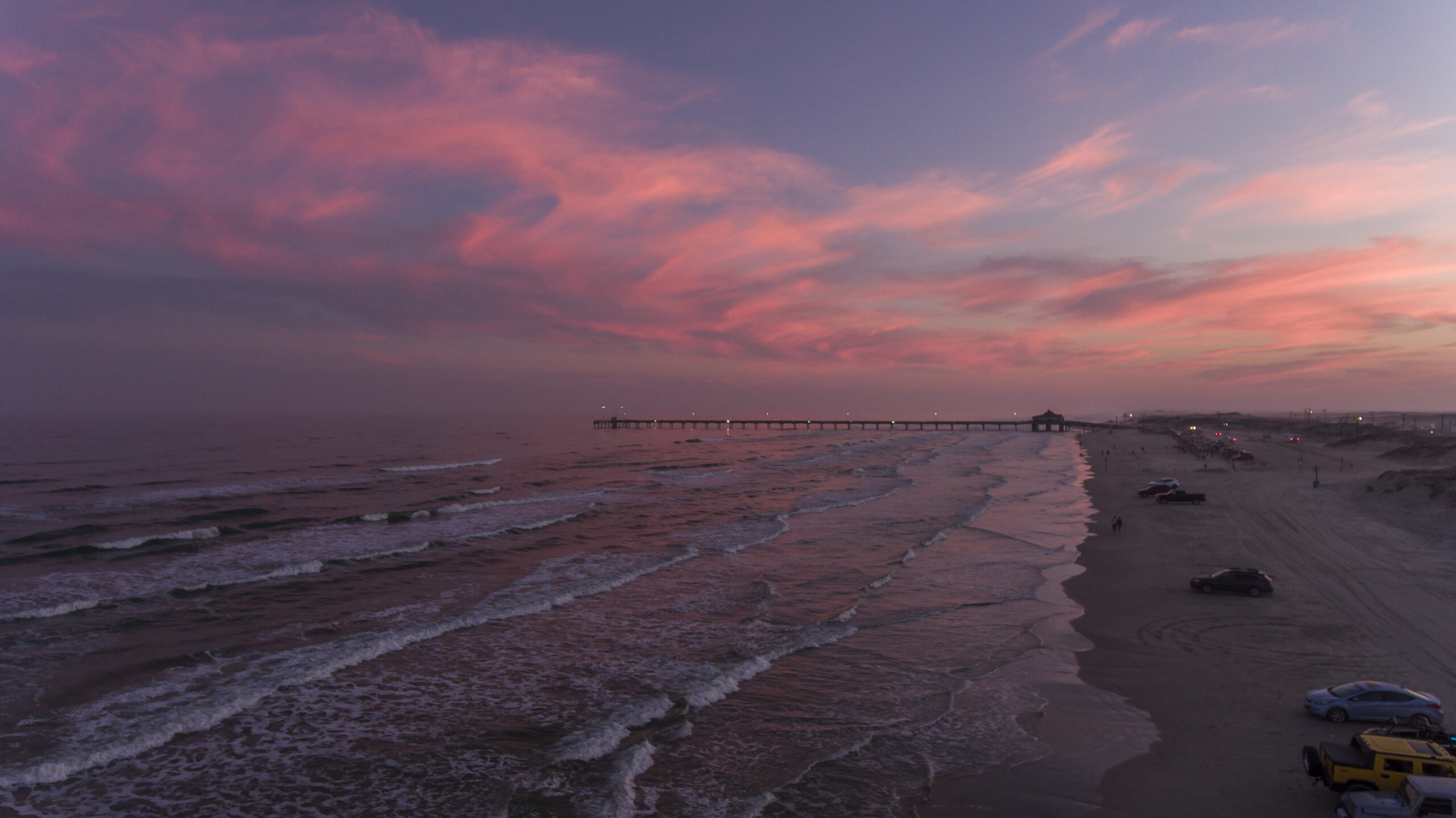 Vehicles line the South Padre Island beach beneath a vivid pink sunset